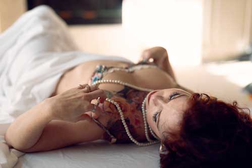 Boudoir photograph of a woman in pearls and lingerie lying on a bed wrapped in white sheets.