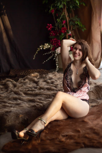 Boudoir photograph of a woman in a pink and black lace lingerie bodysuit and jeweled heels sitting by a bed and laughing.