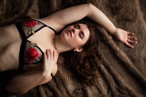 Boudoir photograph of a woman in red and black floral lingerie lying on a bed with her eyes closed. 