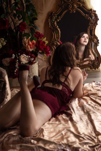 Elegant photograph of a woman in red lingerie lying on a bed and looking past a mirror, surrounded by fine fabrics and flowers.