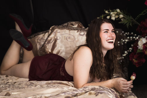 Boudoir photograph of a woman in red lingerie and red high heels lying on a bed laughing and holding a small flower.