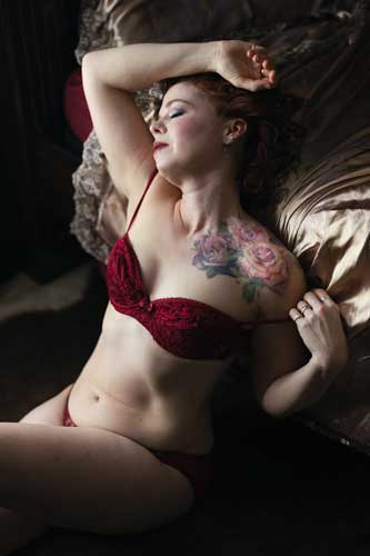 Boudoir photograph of a tatooed mature mom in red lingerie pulling down a bra strap. 