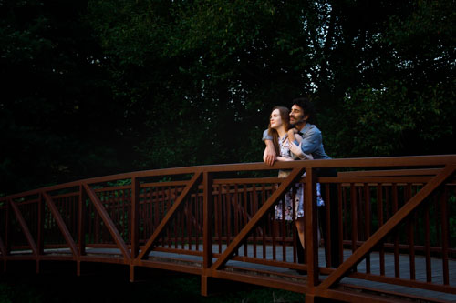 The engaged couple stands on a bridge and looks into the sunset at Shawnee Mission Park.