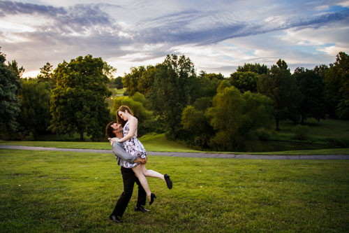 The man lifts his fiancee up in front of a natural, forested setting and striking sky.
