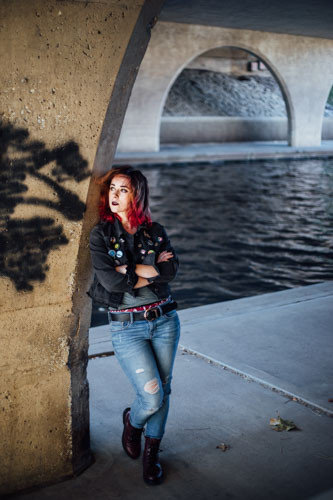 A young woman in punk clothing stands against a graffitied arch under a bridge by the water