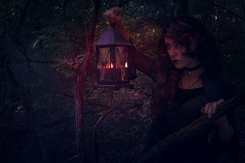 Fantasy photograph of a witchy looking person holding up a lantern while walking through the night forest.