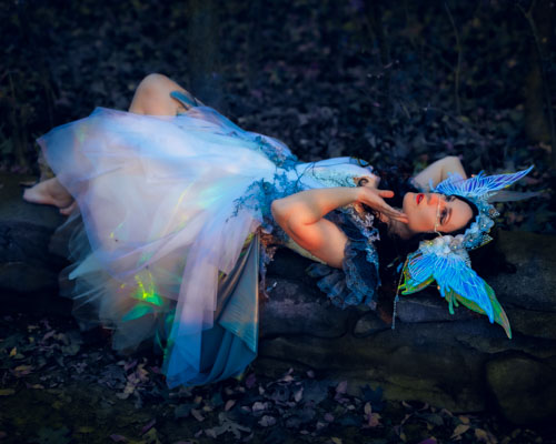 Woman in a fairy costume and fairy headdress lounging on an ancient stone wall in the forest.