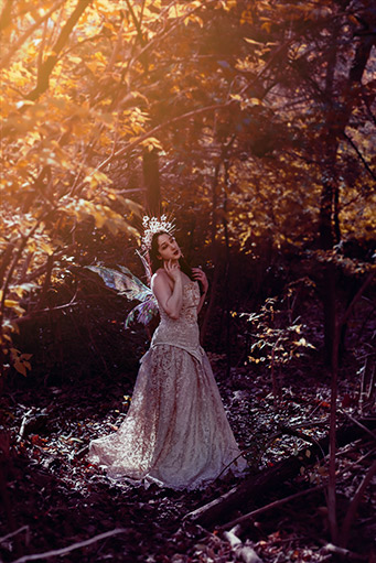 Fine art photograph of a woman costumed in an elegantly trailing gold gown, gold and flower crown, and fairy wings, in the forest.