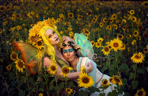 Fantasy photograph of two women in fae costumes and fairy wings with flower headdresses, sitting on the ground and embracing among a field of sunflowers.