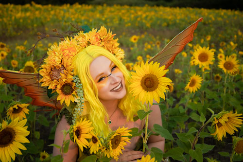 A woman in orange fairy wings and a yellow sunflower headdress smiles next to a big yellow sunflower.