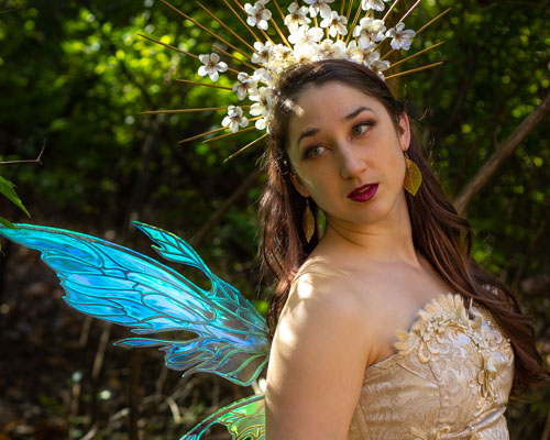 Closeup Photograph of a woman in a gold embellished top, blue and green fairy wings, and a gold and floral headdress in the forest.