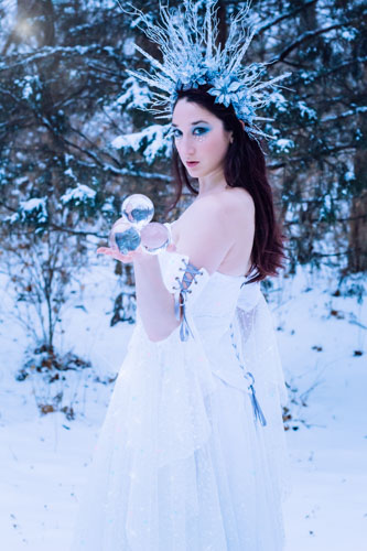Fantasy photograph of a woman in a white glittery gown and elaborate headdress of icicles and silver glittered flowers, holding a pyrimid of clear crystal spheres in the snowy forest.