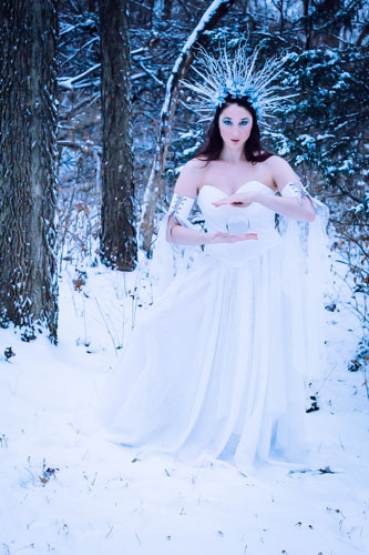 Fantasy photograph of a woman in a white glittery gown and elaborate headdress of icicles and silver glittered flowers, holding a clear crystal orb in the snowy forest.