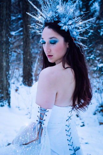 Fantasy photograph of a woman in a white glittery gown and elaborate headdress of icicles and silver glittered flowers, in the snowy forest.