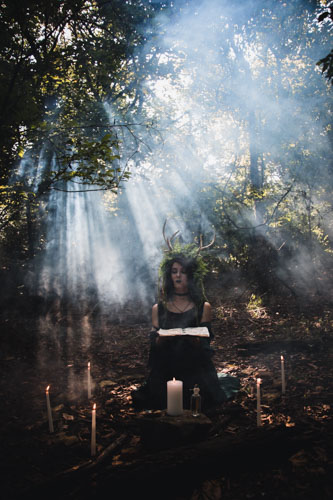 Fantasy photograph of a non-binary person in a black dress and headdress of leaves, vines, and antlers, kneeling in the forest surrounded by candles, holding a spellbook, with the sunlight streaming through the fog behind.