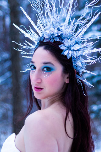 Fantasy photograph closeup of a woman in a white glittery gown and elaborate headdress of icicles and silver glittered flowers, in the snowy forest.