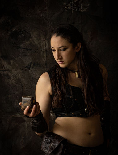 Fine art photograph of a woman in a black leather neo-tribal style crop top and leather arm bracers looking at a compass in front of a painterly backdrop.