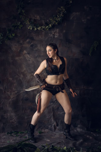 Fine art photograph of a woman in post-apocalyptic style outfit of black leather boots, crop top, layered belts, and brown leather arm bracers holding a machete and looking behind her.
