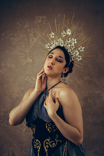 Fine art photograph of a woman costumed in a blue gown and gold floral headdress, in front of a painterly tan backdrop.