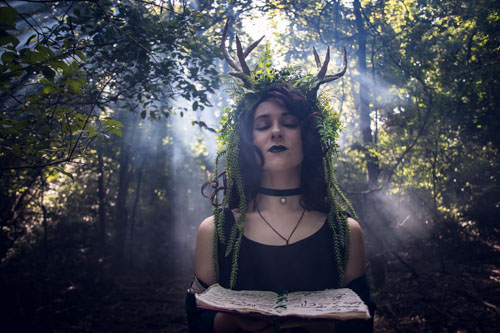 Fantasy photograph of a non-binary person in a black dress and headdress of leaves, vines, and antlers, in the forest holding a spellbook, with the sunlight streaming through the fog behind.