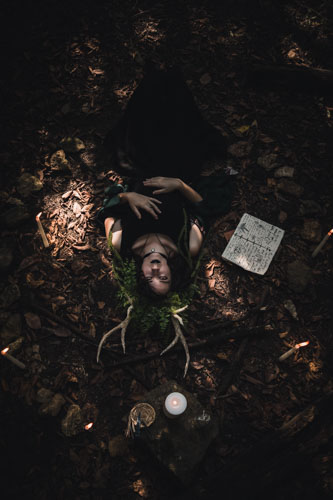 Fantasy photograph of a non-binary person in a black dress and headdress of leaves, vines, and antlers, lying on the ground upside down looking up at the camera, surrounded by candles, stones, and spellbooks.