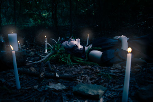 Fantasy photograph of a non-binary person in a black dress and headdress of leaves, vines, and antlers lying on the ground and looking up at the sky, surrounded by candles.