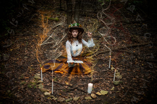 Fantasy photograph of a woman in an orange skirt, white lace shirt, and brown witch hat holding a glowing spellbook and appearing to use her hands to cast a spell, surrounded by a circle of stones and candles in the woods. Colored smoke rises from teh candles, and arcane symbols encircle her.