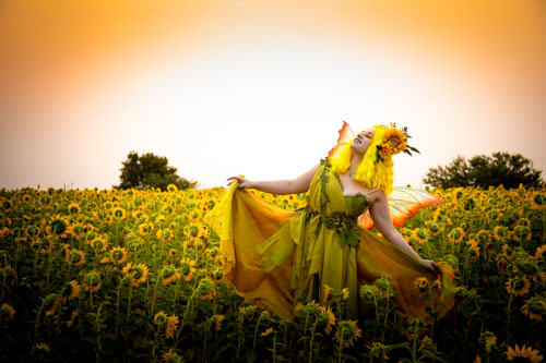 Fantasy photograph of a woman in a green and yellow fairy costume and orange fairy wings dancing in a field of sunflowers.