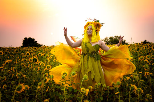 Fantasy photograph of a woman in a green and yellow fairy costume and orange fairy wings twirling her skirt in a field of sunflowers.