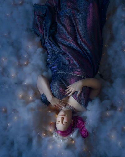 Woman in a blue glittery dress and star crown lying in a bed of clouds and stars.