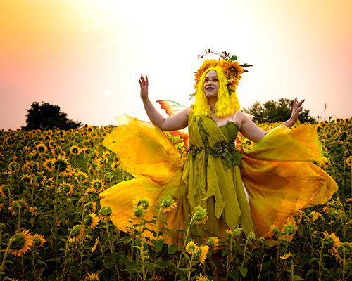 Photograph of a woman in an elegant yellow fairy costume dancing in a sunflower field