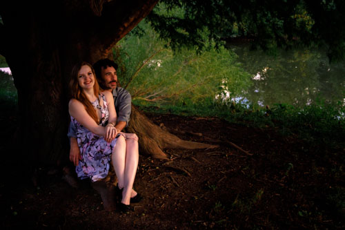 The engaged couple sits underneath a tree in the dusk sunlight.