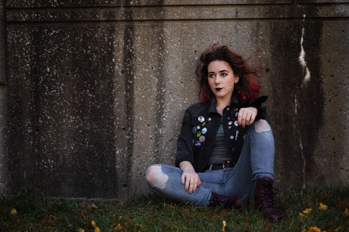 A young woman in a jean jacket and ripped jeans sits against a weathered stone wall