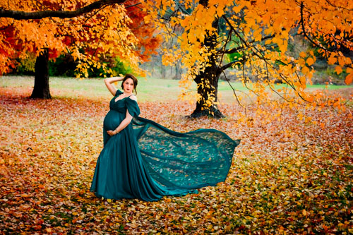 A pregnant woman in a teal dress blown by the wind stands under a tree with orange and red leaves