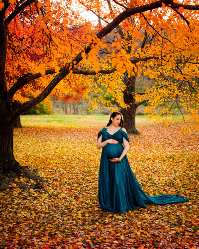 A pregnant woman in a teal dress holds her belly in a park in autumn, surrounded by orange and red leaves