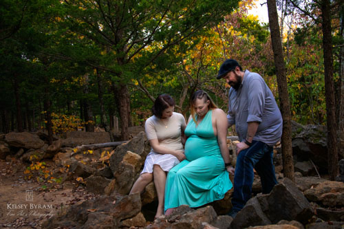 A pregnant woman in a seafoam green dress sits on boulders and looks at her belly, while her partners stand by her