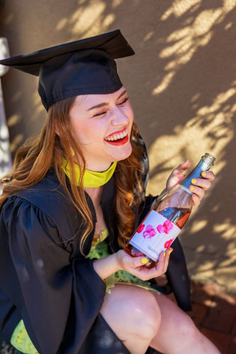 A woman in a black graduation gown and yellow dress holds a bottle of champagne and laughs