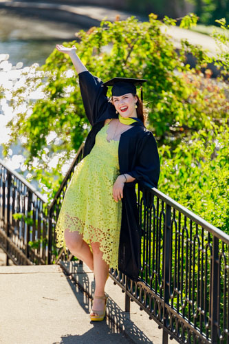 A woman in a black graduation hat and gown and yellow dress raises her arm in celebration while leaning against a railing in front of a river