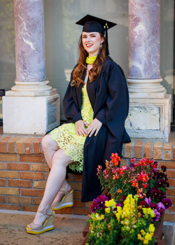 A woman in a black graduation cap and gown and lemon yellow dress sits on a brick wall between pillars