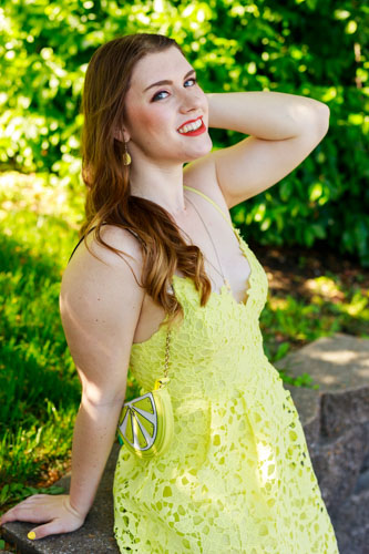 A young woman in a yellow dress and lemon purse leans against a stone wall in front of grass and bushes