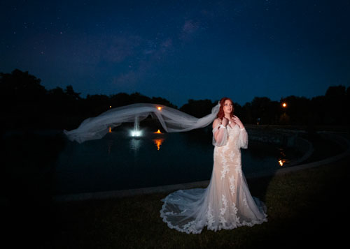 A bride in a cream dress stands in front of a lake at night, with her veil flowing behind her