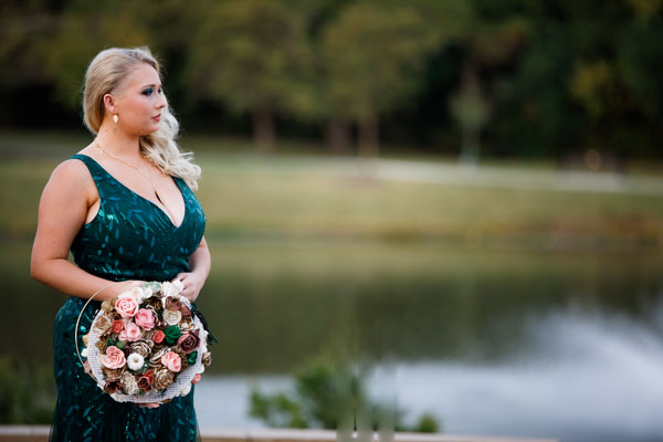 A woman in an emerald green dress holds flowers in front of a lake and forest
