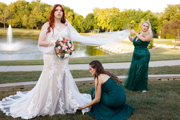 The bride looks thoughtful while the maid of honor holds her veil and a bridesmaid adjusts the hem of her dress