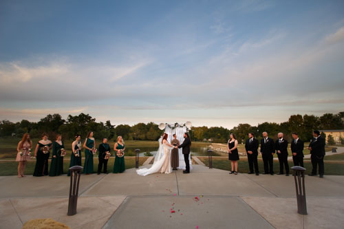 A wide view of the entire wedding party standing around the bride and groom holding hands in front of the officiant and wedding arch, in front of a lake and forest
