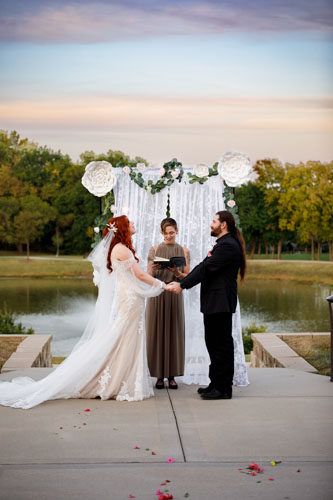 The bride and groom hold hands in front of the officiant and floral wedding arch in front of a lake and trees