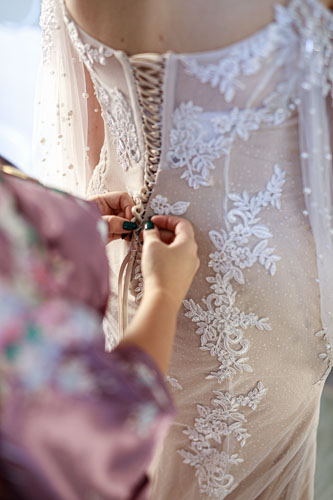 A view over the shoulder of the maid fo honor as she laces up the back of a cream and white lace wedding gown